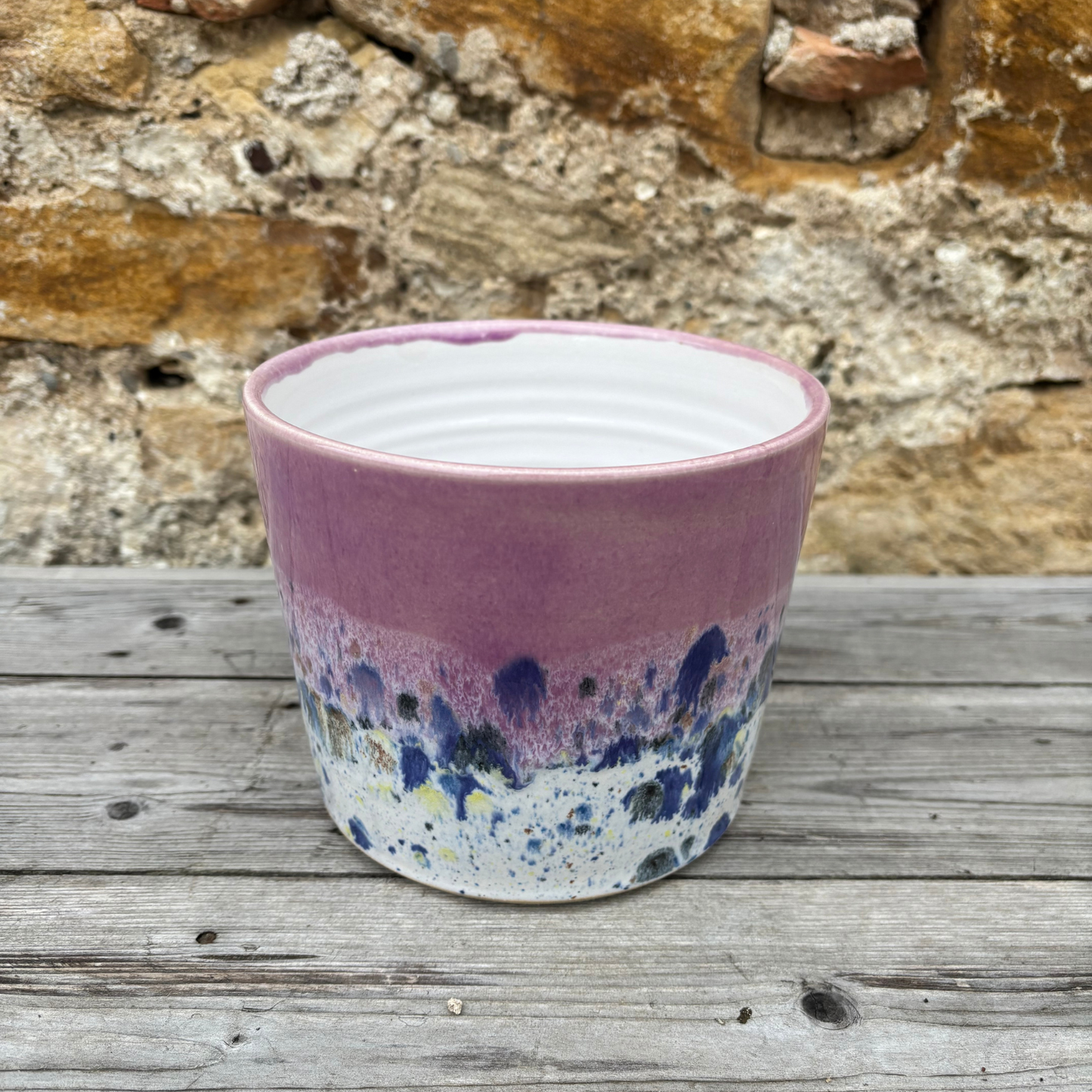 Ceramic pot with a purple and blue speckled design on a wooden surface with a stone wall background