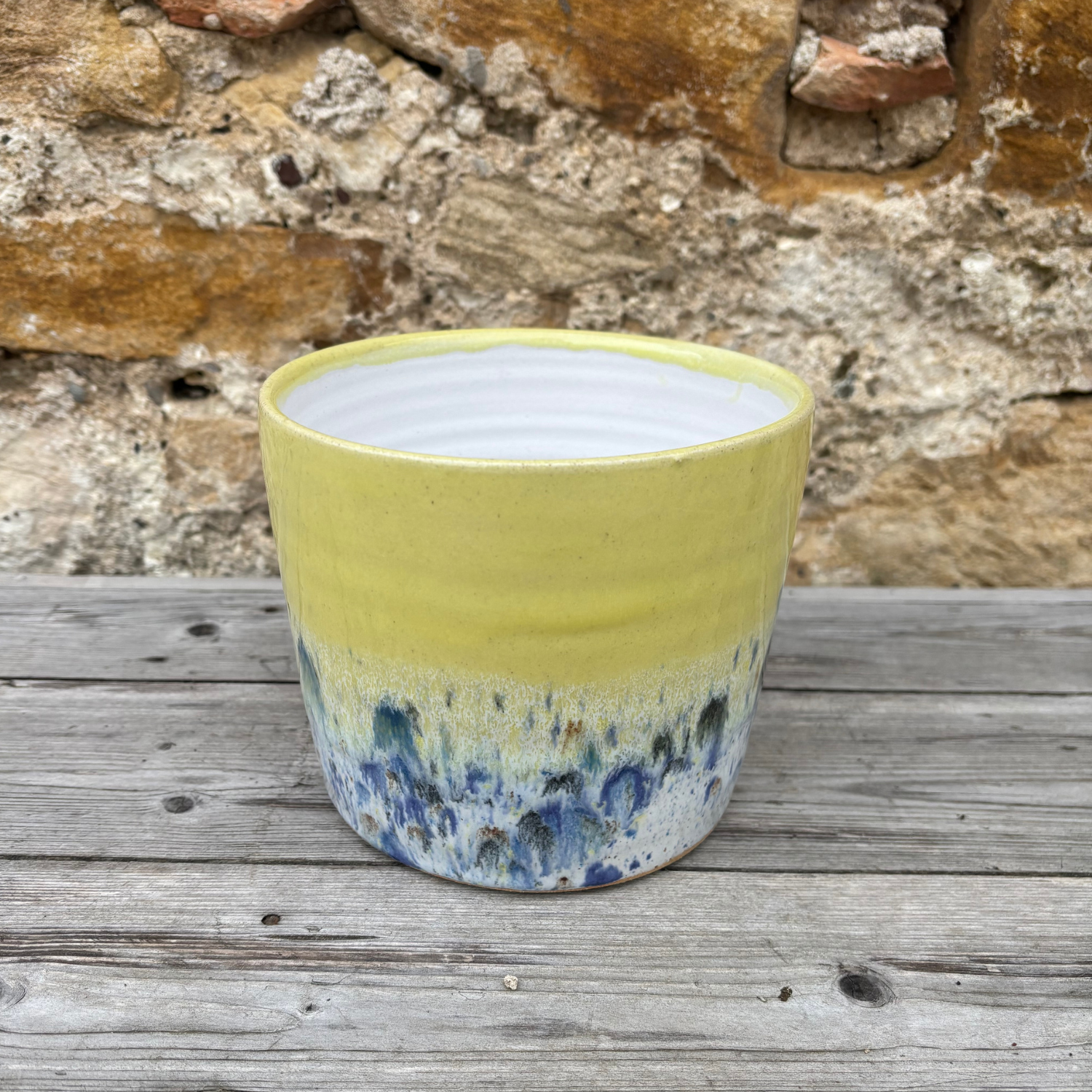 Yellow ceramic pot with blue speckled design on a wooden surface with stone wall background