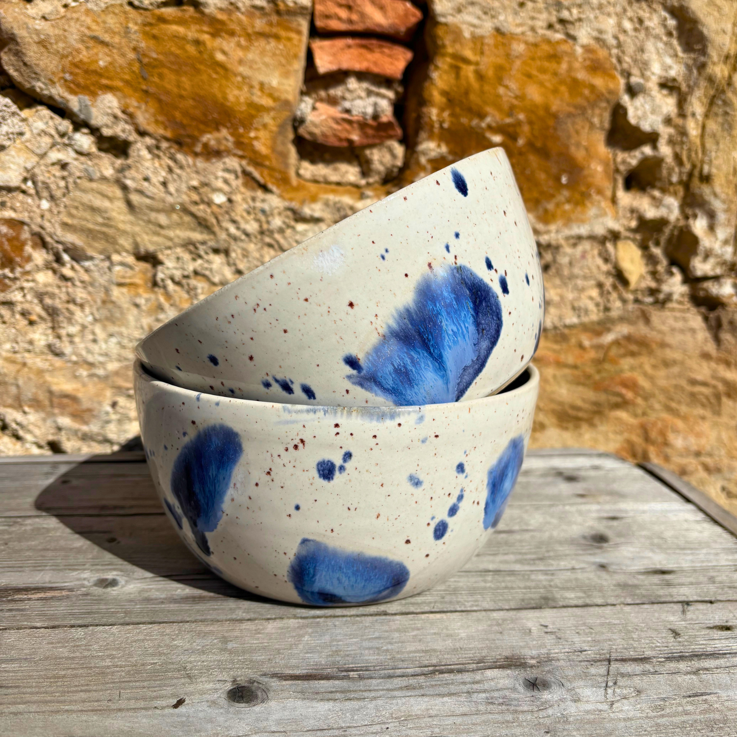 Speckled ceramic bowls with blue splashes on a wooden surface against a stone wall.