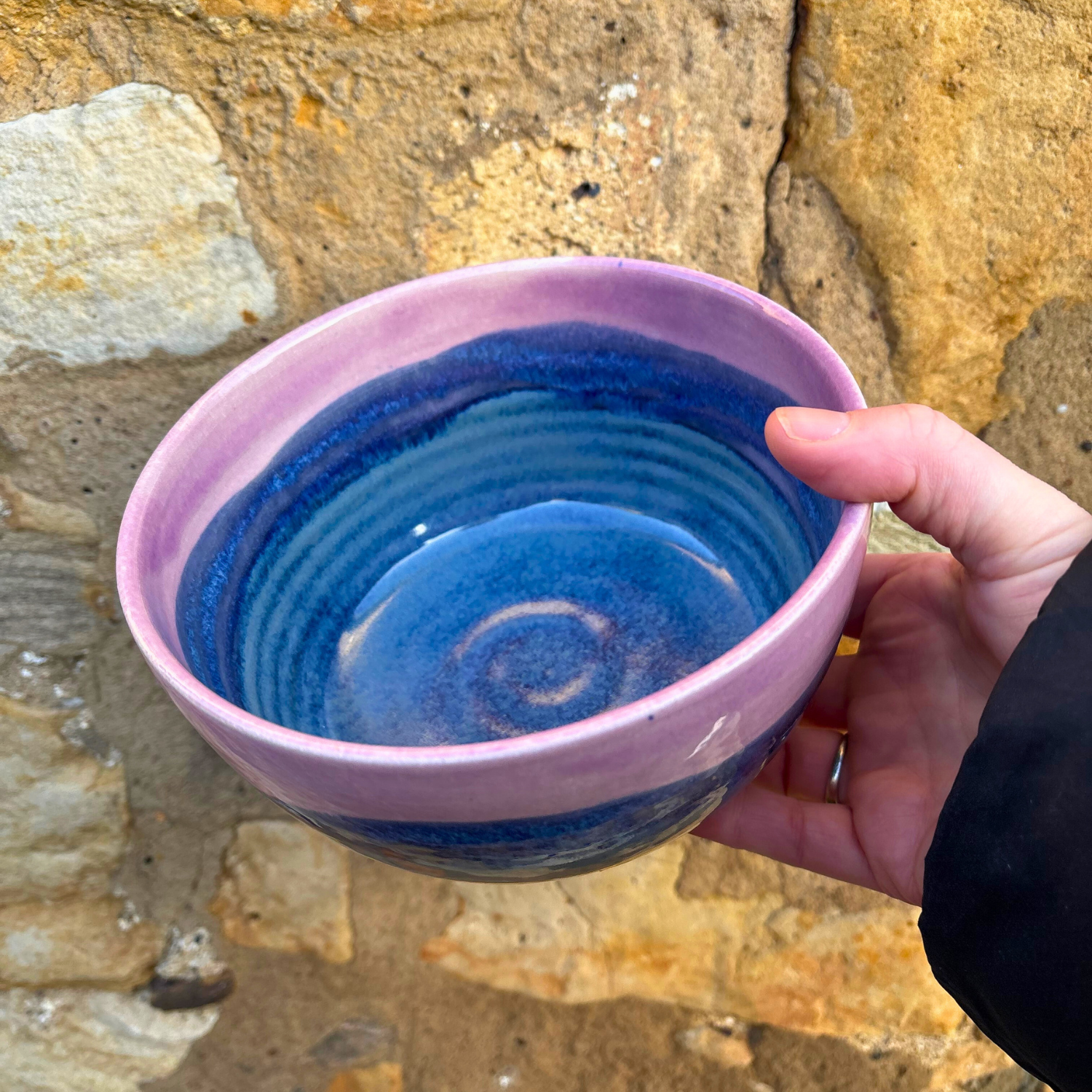 Hand holding a blue ceramic bowl with a pink rim against a stone wall background