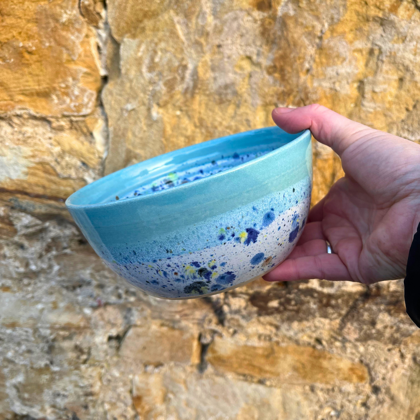 Hand holding a blue ceramic bowl with speckled design against a stone background