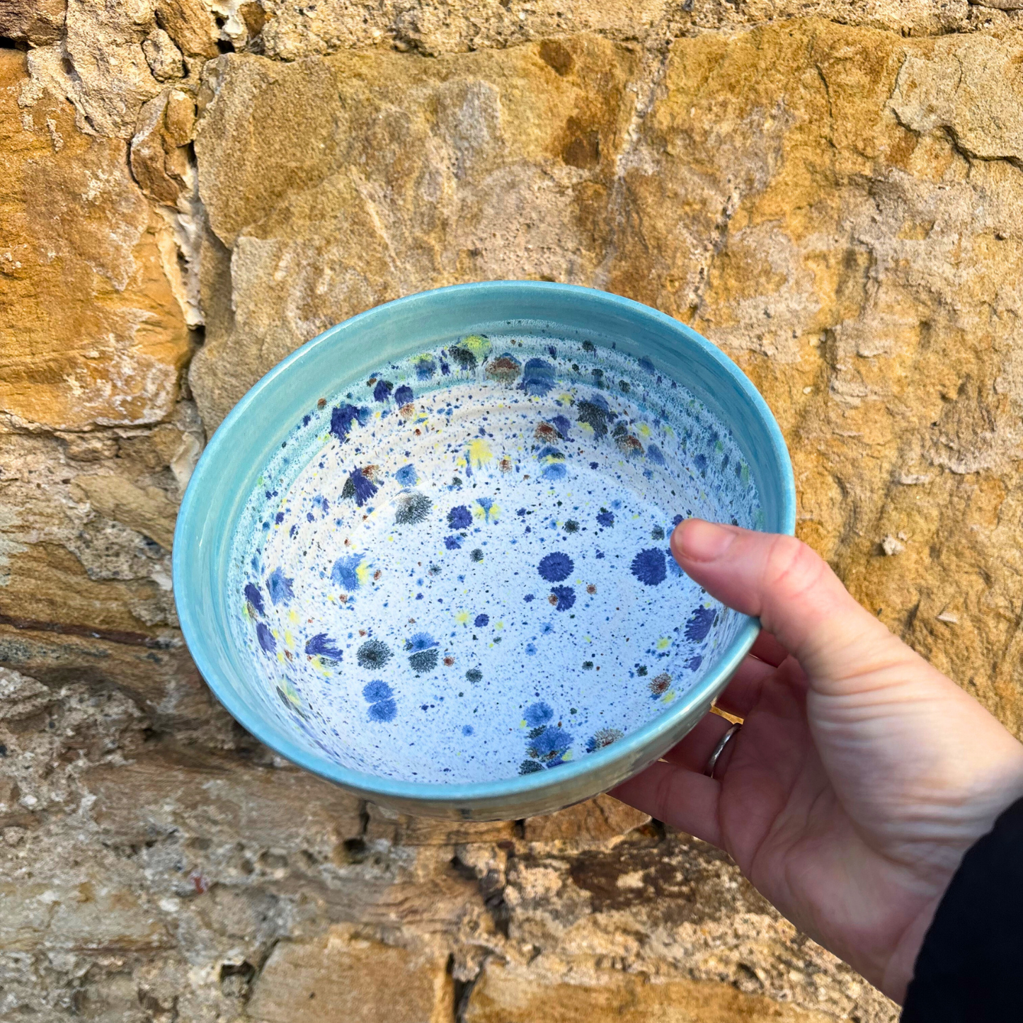 Hand holding a blue ceramic bowl with speckled design against a stone wall background