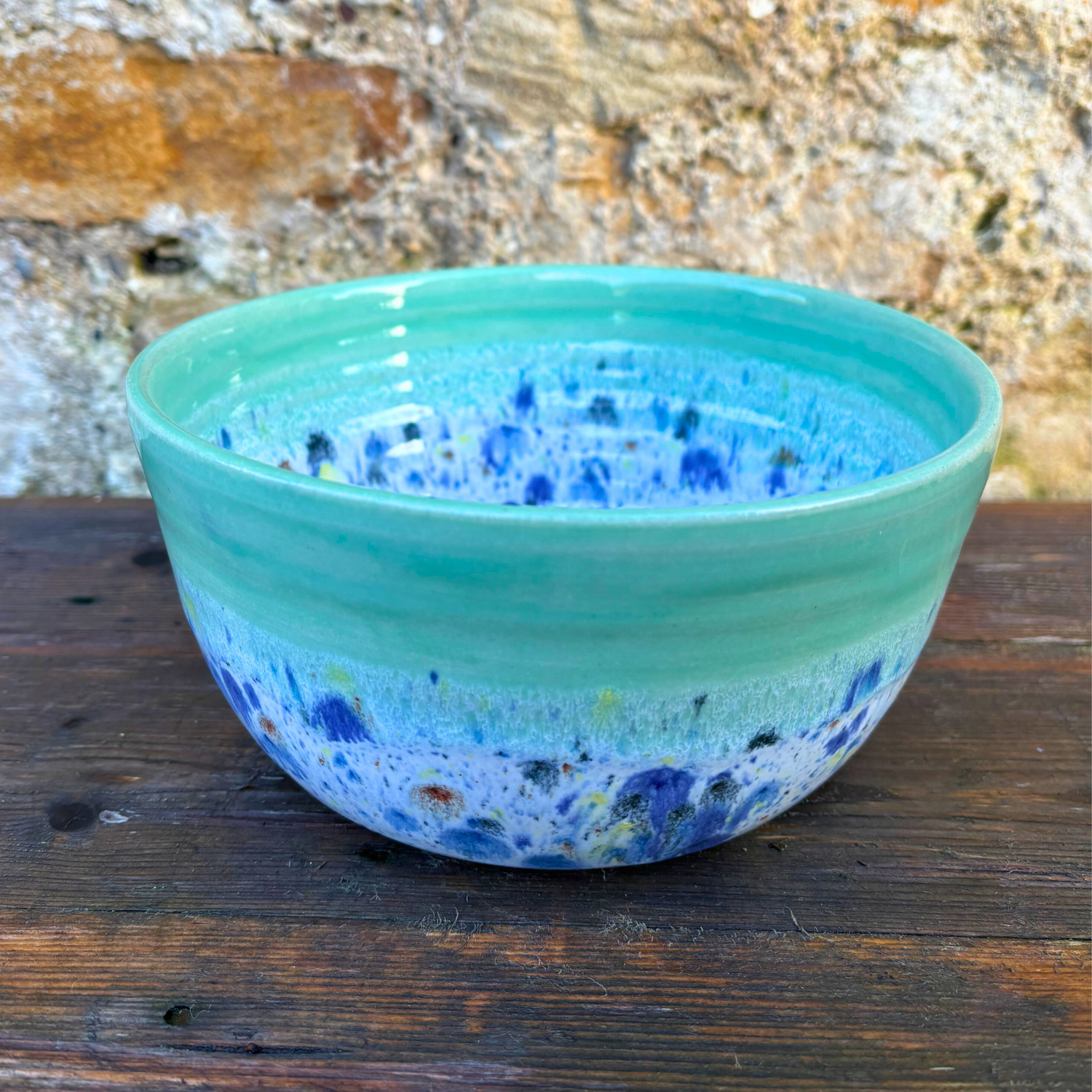 Turquoise ceramic bowl with blue speckles on a wooden surface with a stone wall background