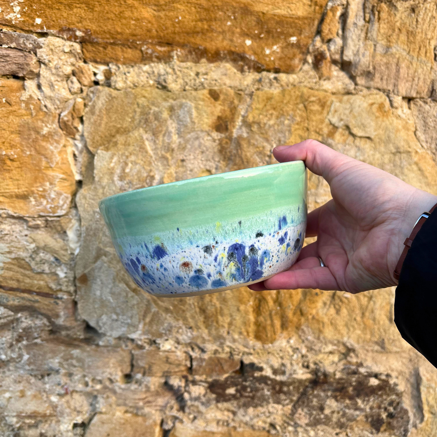 Hand holding a green ceramic bowl with blue speckles against a stone wall.