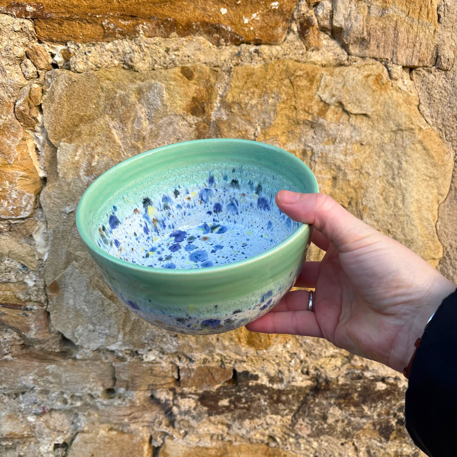 Hand holding a green ceramic bowl with blue speckles against a stone wall background