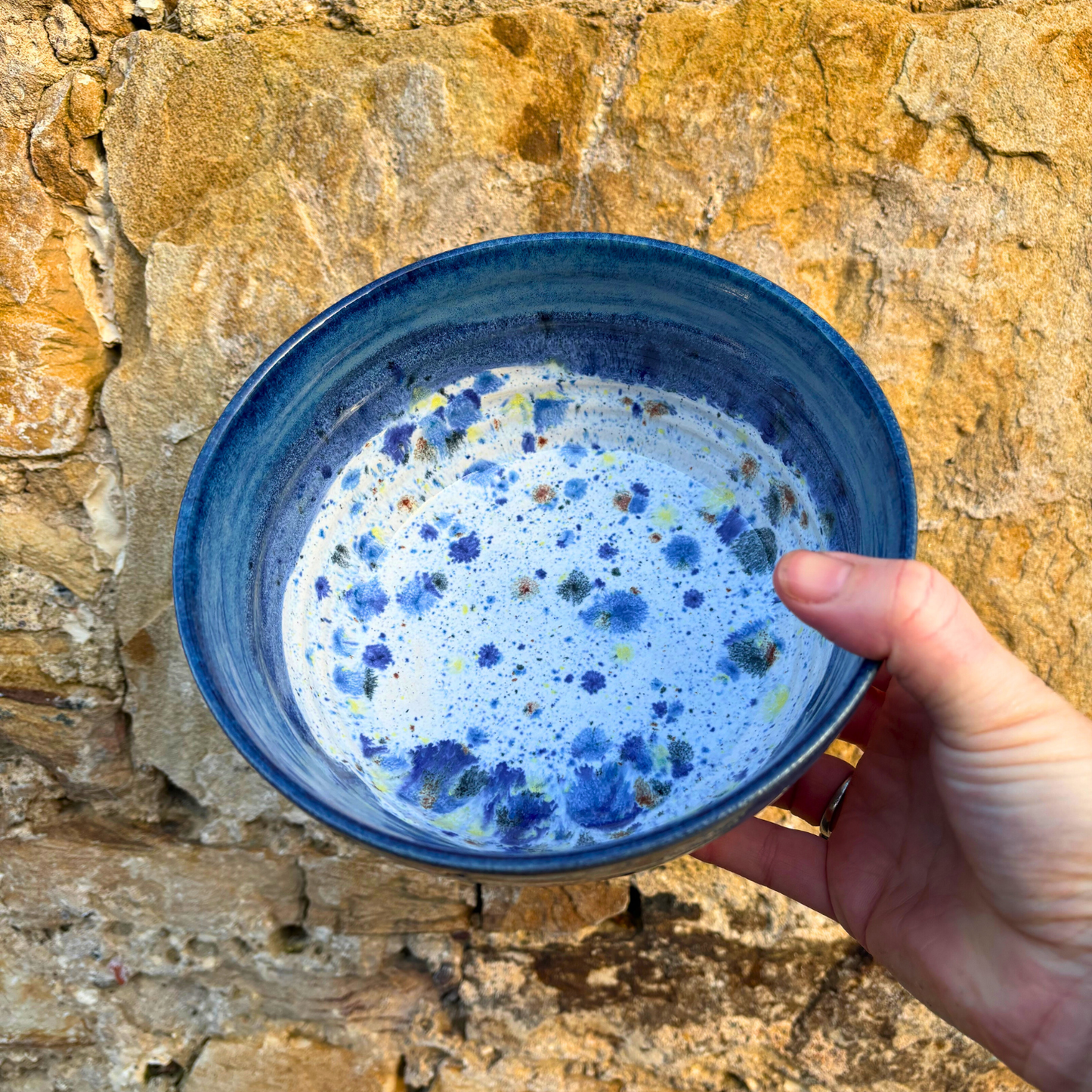 Hand holding a blue ceramic bowl with speckled design against a stone wall background