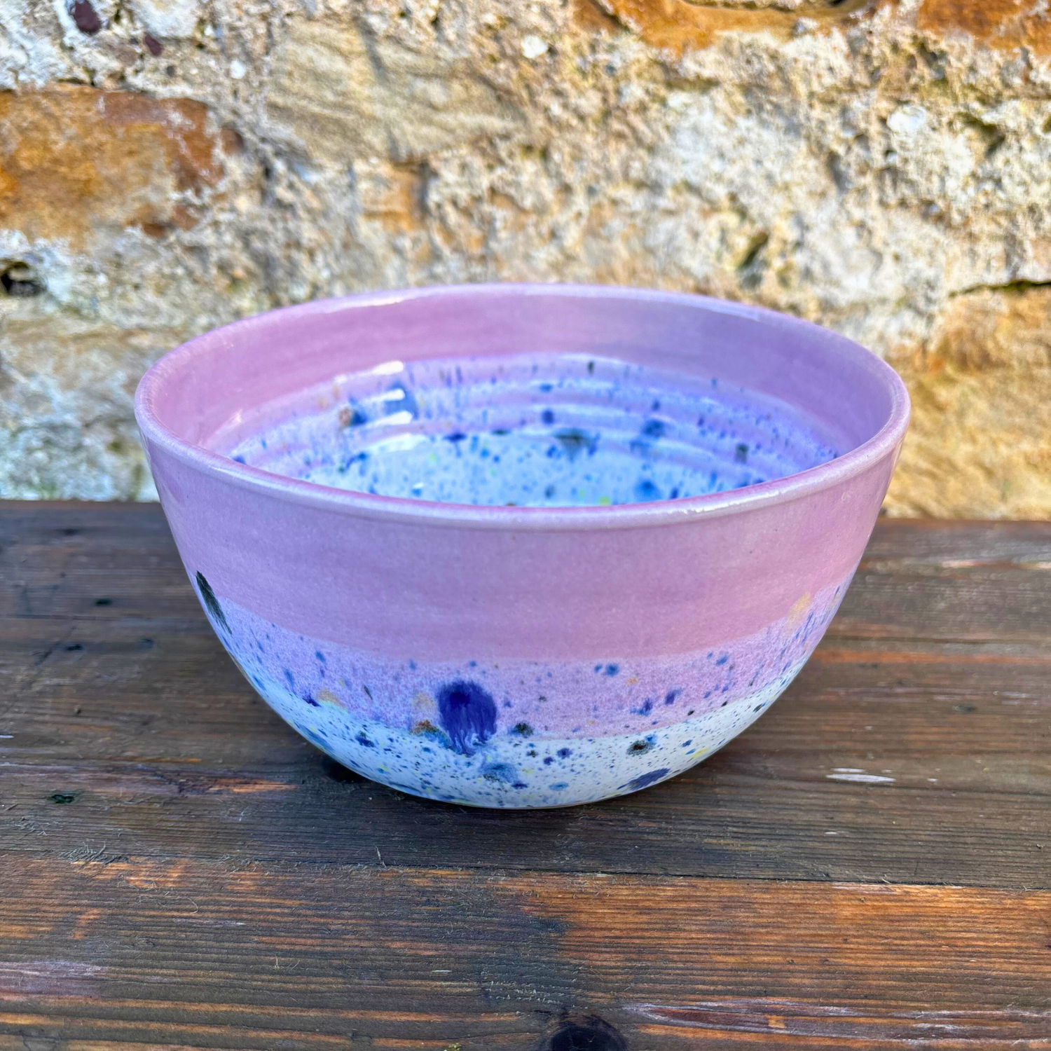 Pink ceramic bowl with blue speckles on a wooden surface with a stone wall background