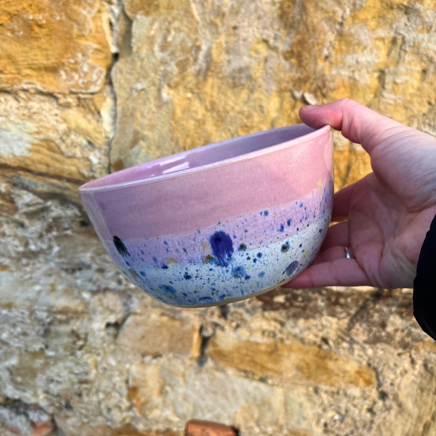 Hand holding a ceramic bowl with pink and blue speckled design against a stone wall background