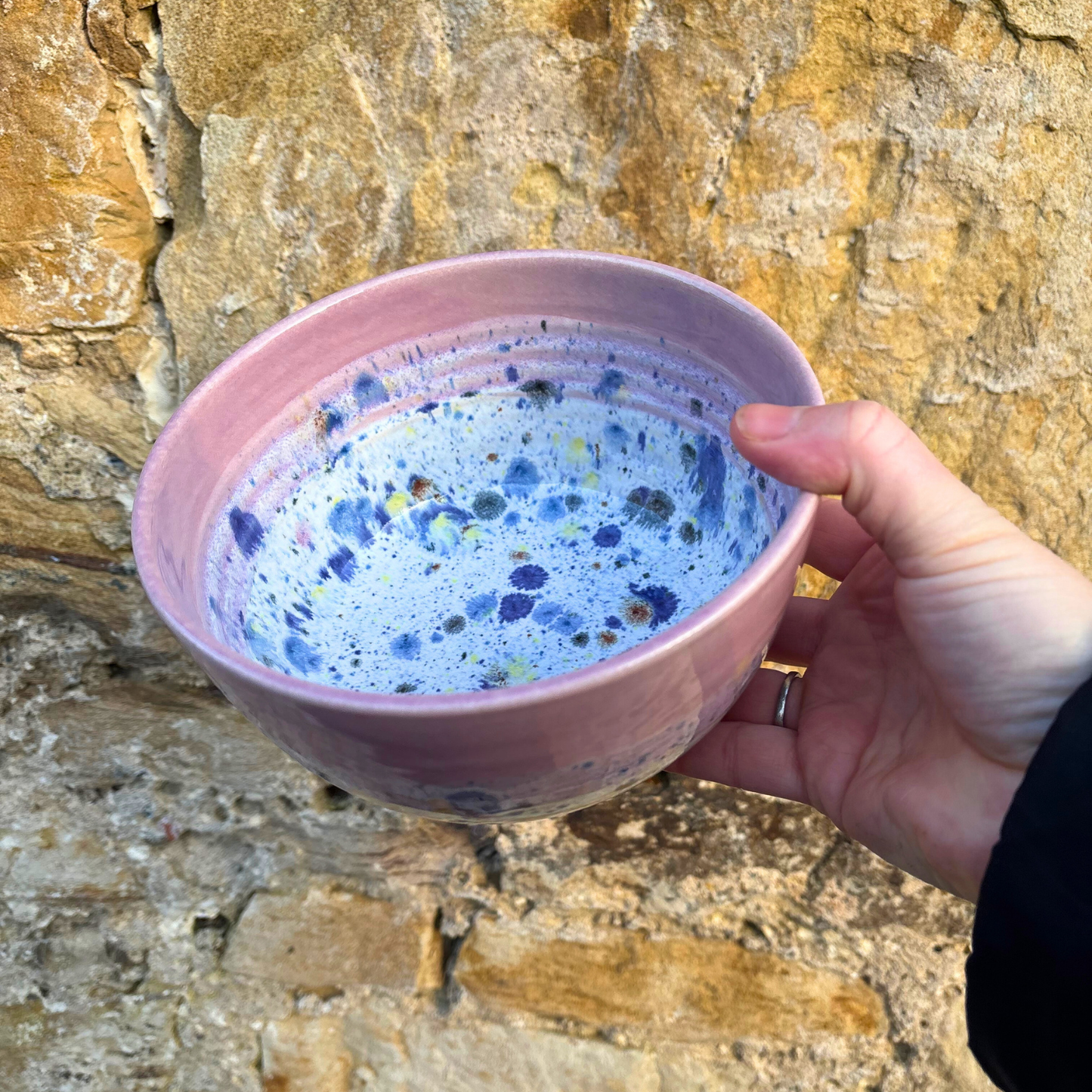Hand holding a pink ceramic bowl with blue speckles against a stone wall background