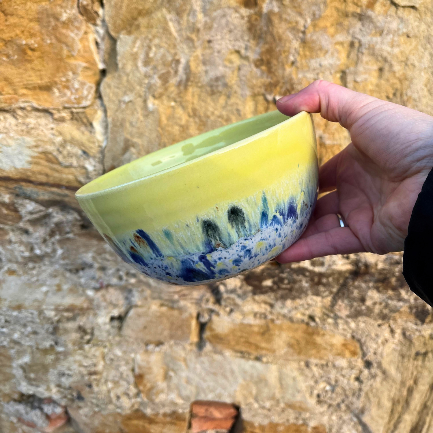 Hand holding a yellow ceramic bowl with blue and white design against a stone wall background