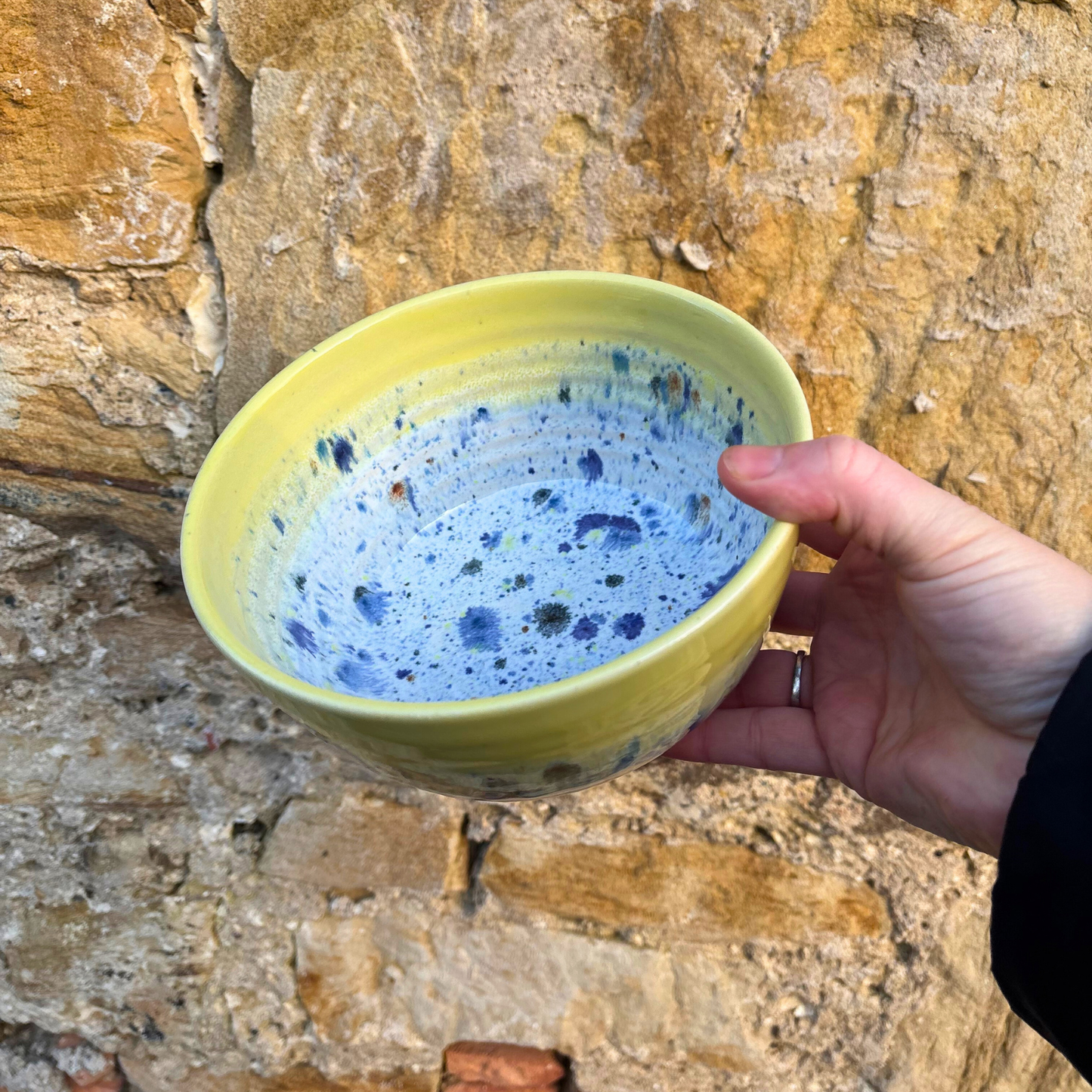 Hand holding a yellow ceramic bowl with blue speckles against a stone wall background