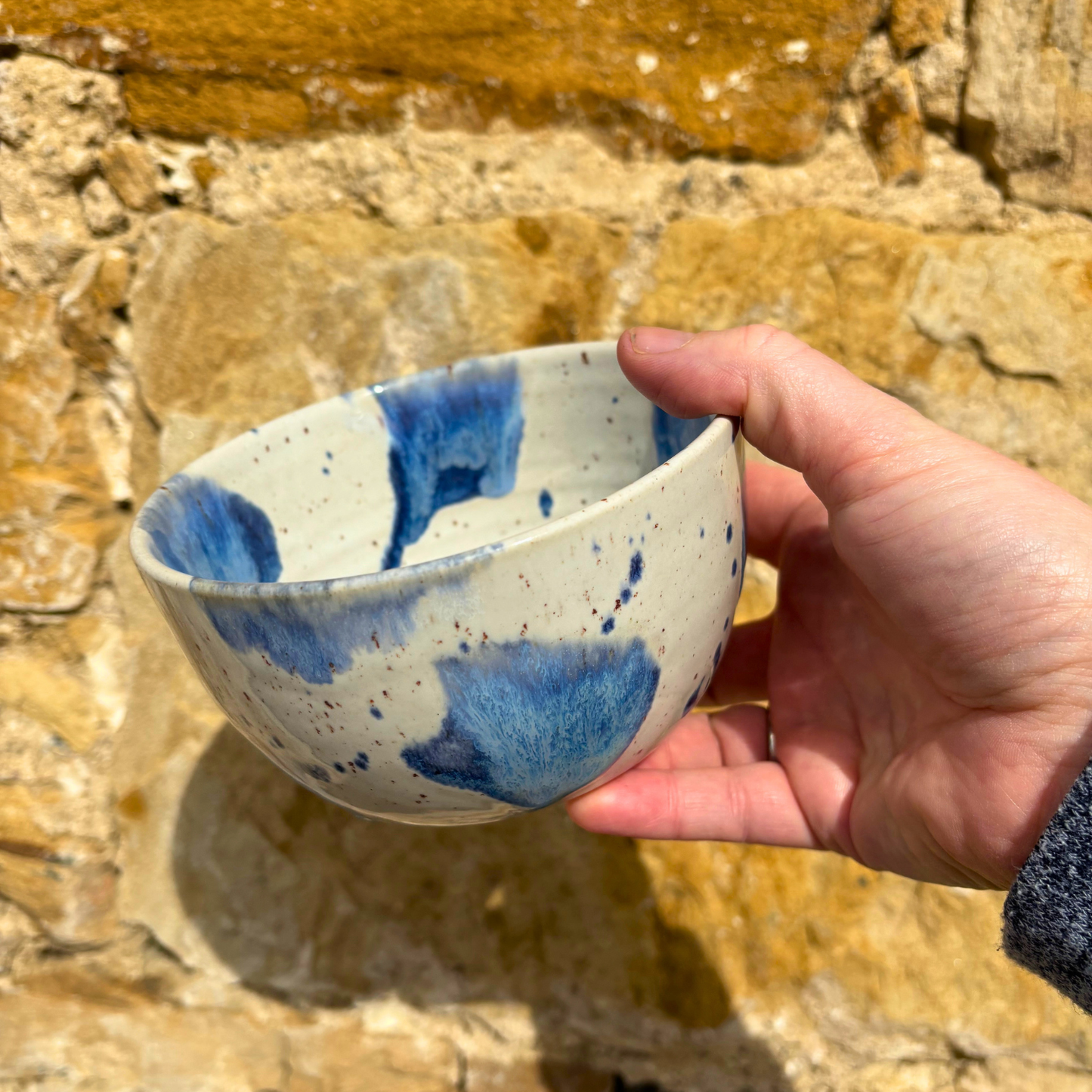 Hand holding a ceramic bowl with blue speckles against a stone wall background
