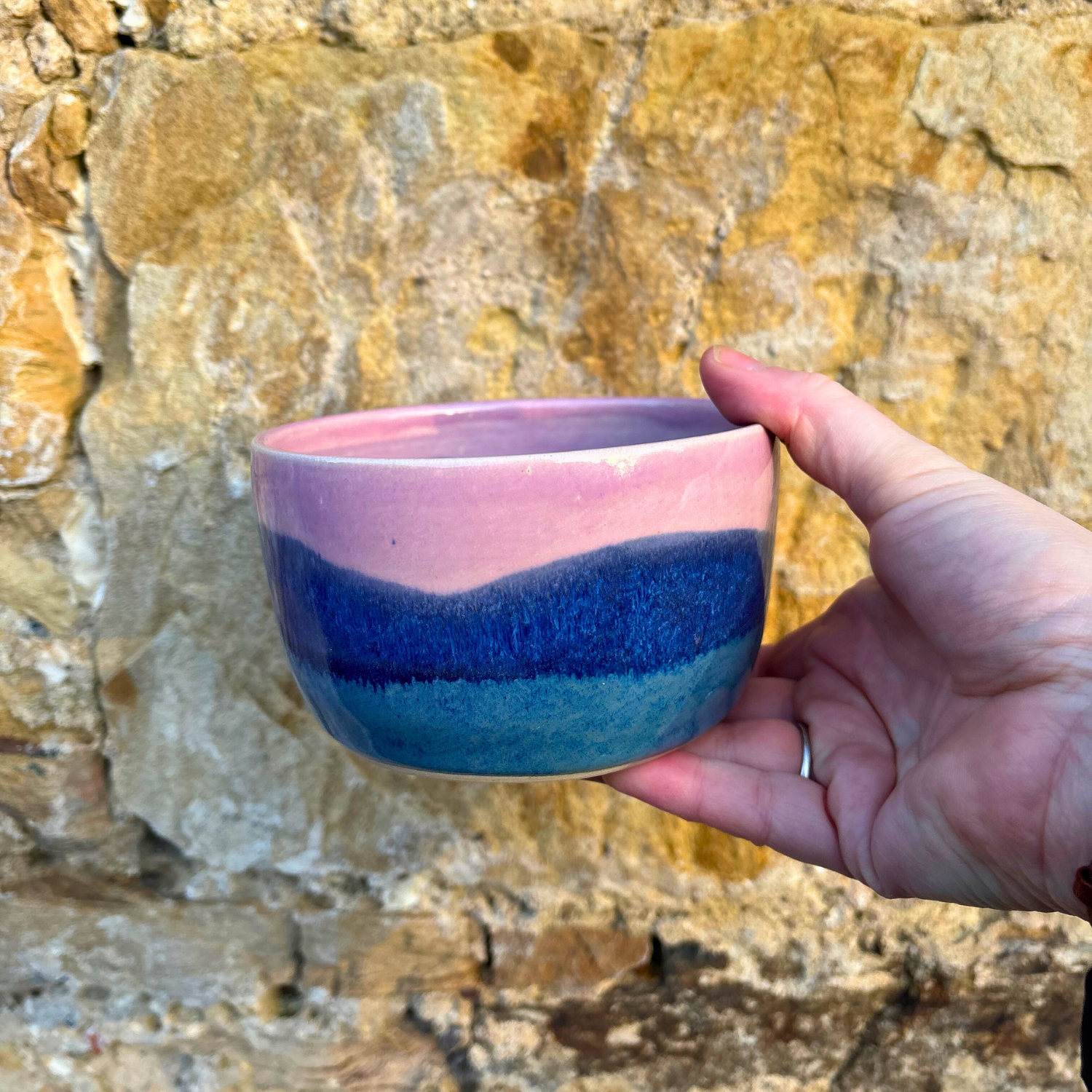 Hand holding a ceramic cup with pink and blue layers against a stone wall background