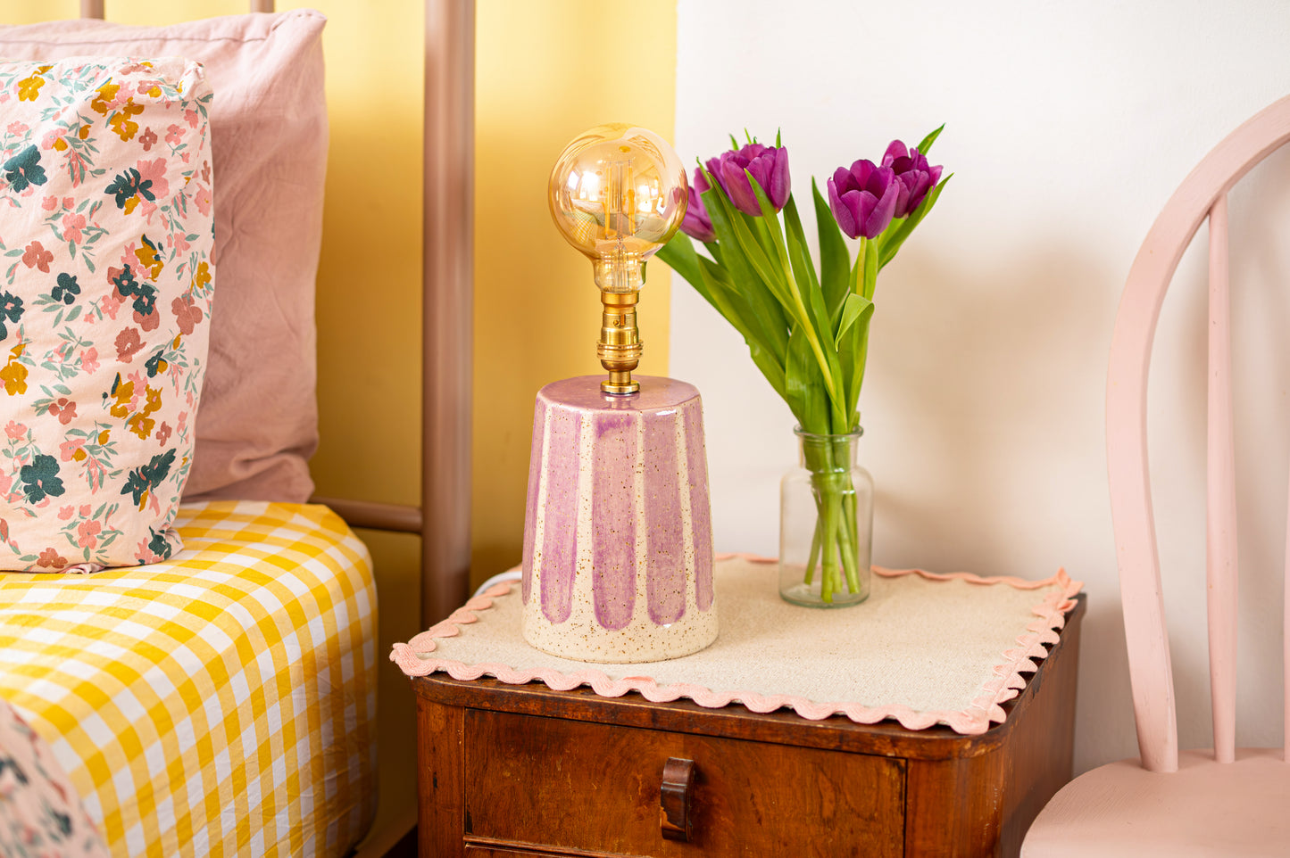 Small wooden table with a pink lamp and purple tulips on a bed with floral and checkered bedding.
