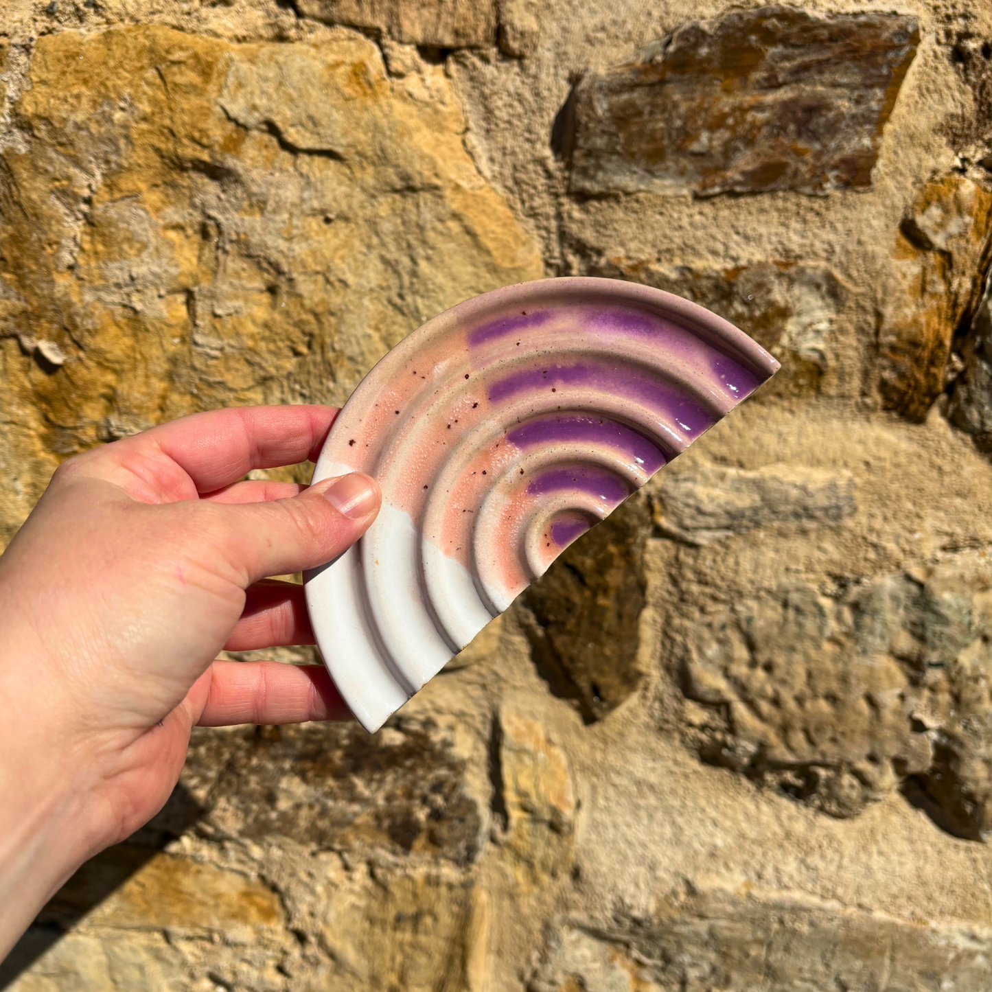 Hand holding a ceramic soap dish with purple and pink concentric circles against a stone wall background