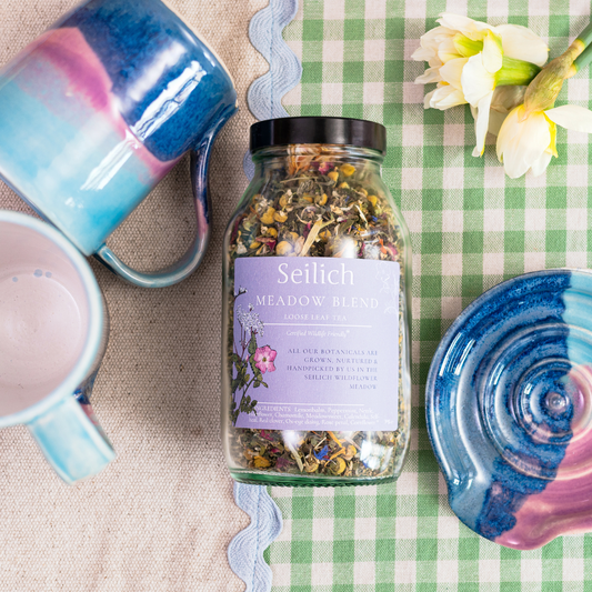 Seilich Meadow Blend tea jar on a checkered tablecloth with sunrise landscape mugs and flowers.