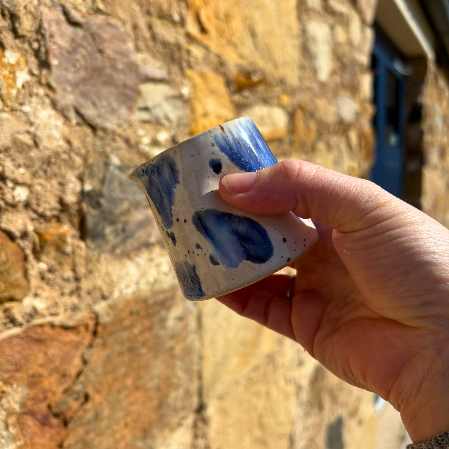 Hand holding a small ceramic cup with blue speckles against a stone wall background