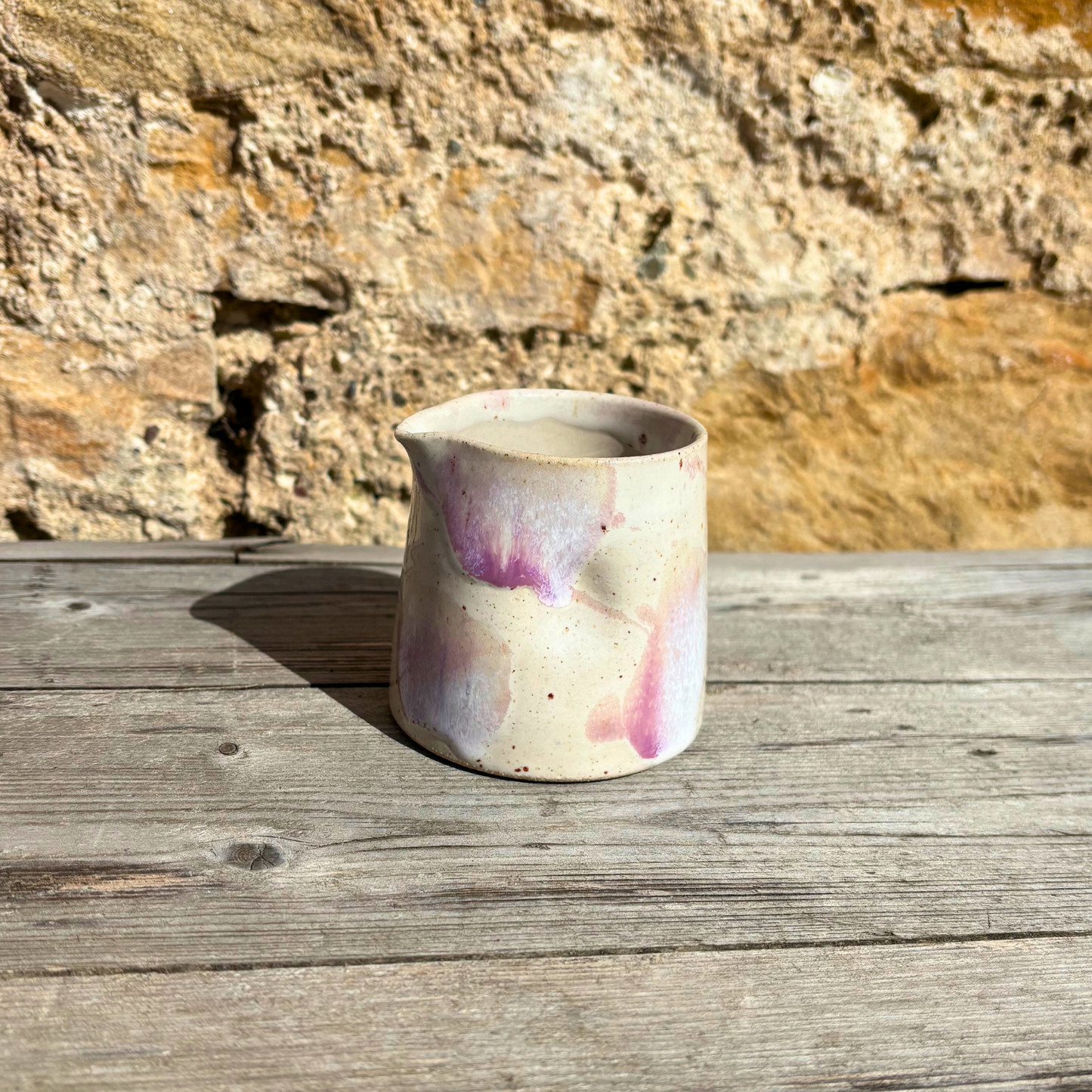 Ceramic pitcher with purple and pink glaze on a wooden surface with a stone wall background