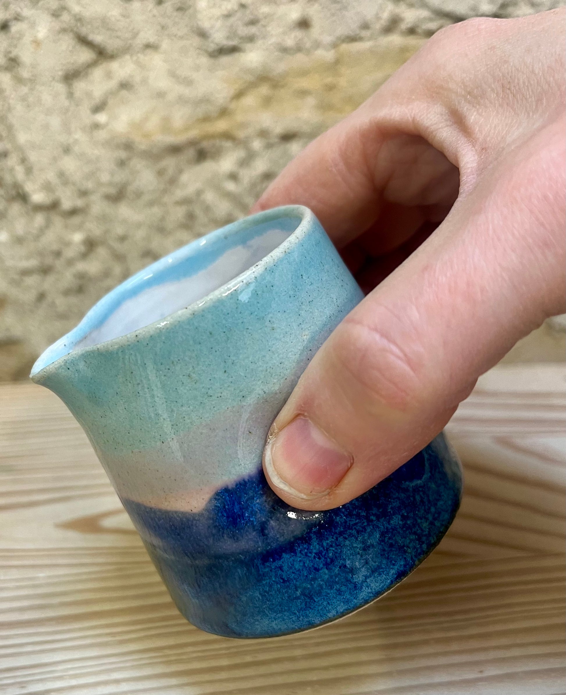 Hand holding a small ceramic pitcher with blue and white glaze on a wooden surface.