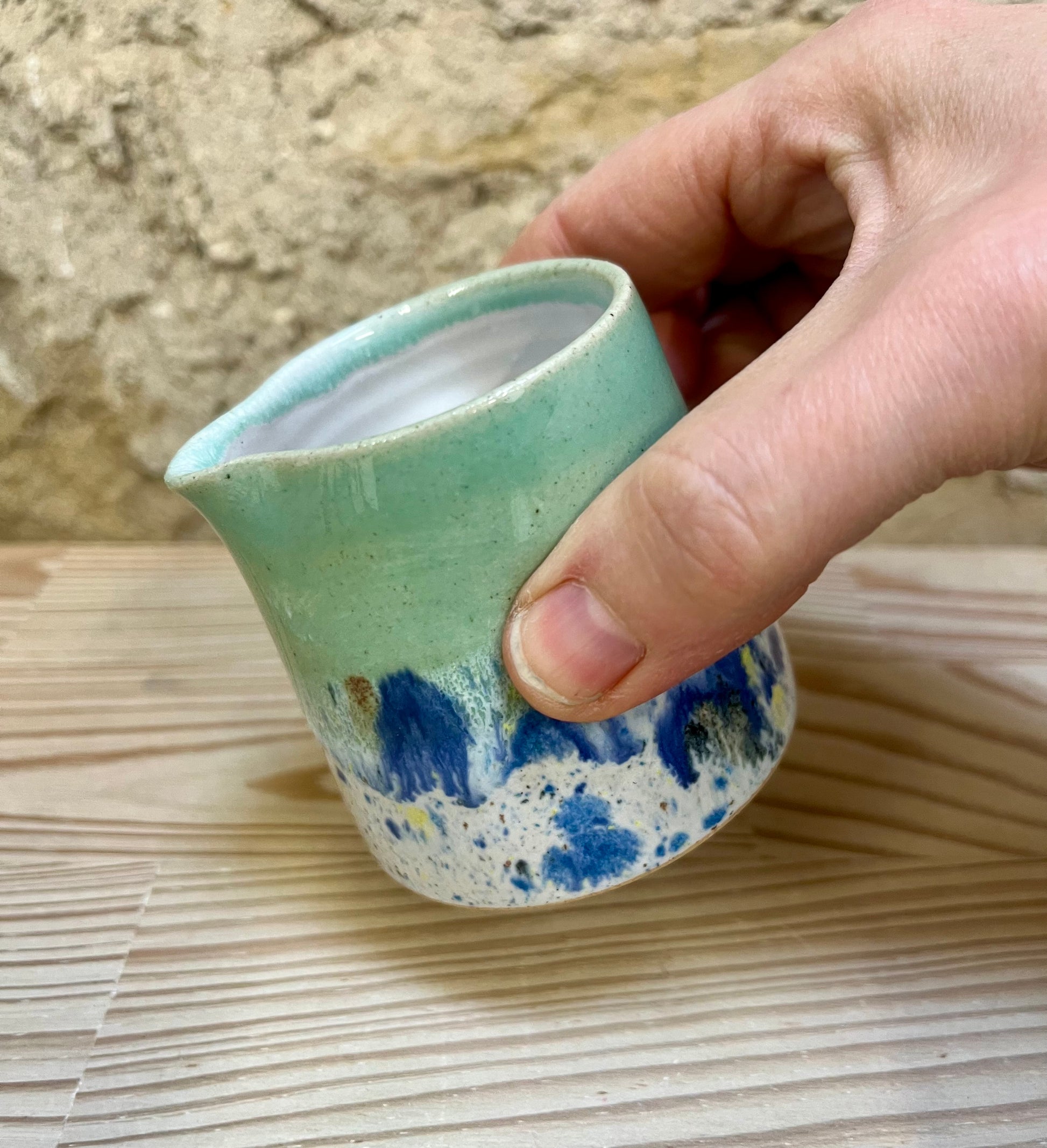 Hand holding a small ceramic pitcher with green and blue speckled design on a woven mat.