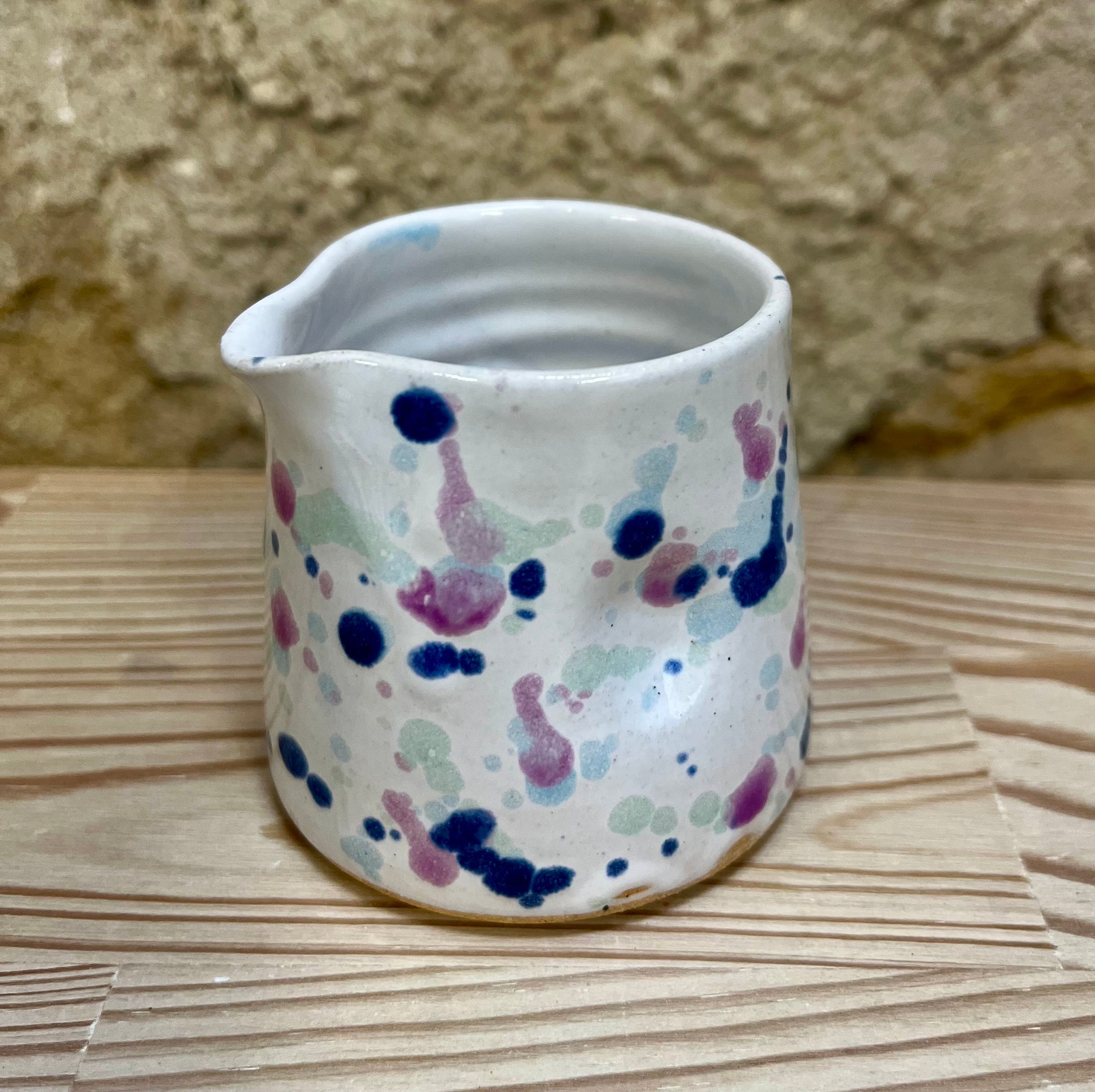 Small ceramic pitcher with multicolored speckles on a wooden surface with a stone wall background
