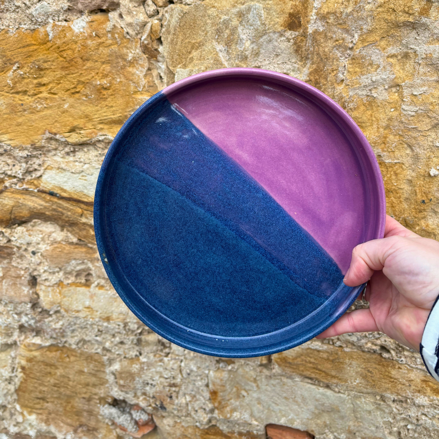 Hand holding a two-toned purple and blue ceramic tray against a stone background