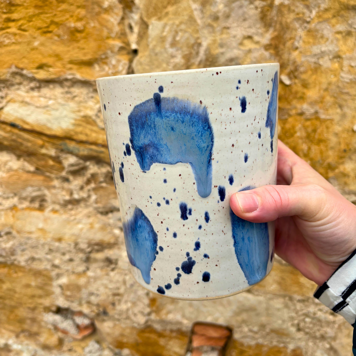 Hand holding a ceramic utensil pot with blue splatter design against a stone background