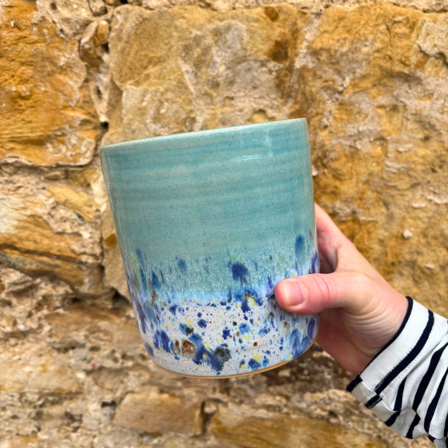 Hand holding a blue ceramic utensil pot with speckled design against a stone wall background