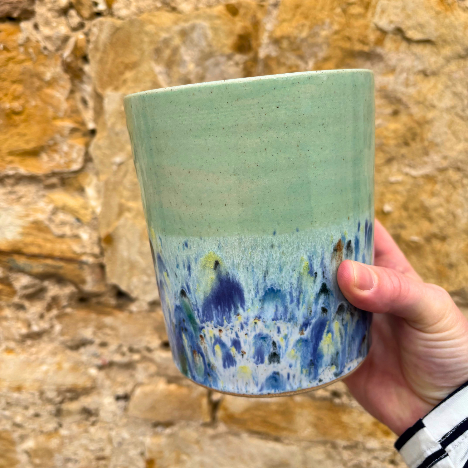 Hand holding a ceramic utensil pot with a gradient design against a stone wall background