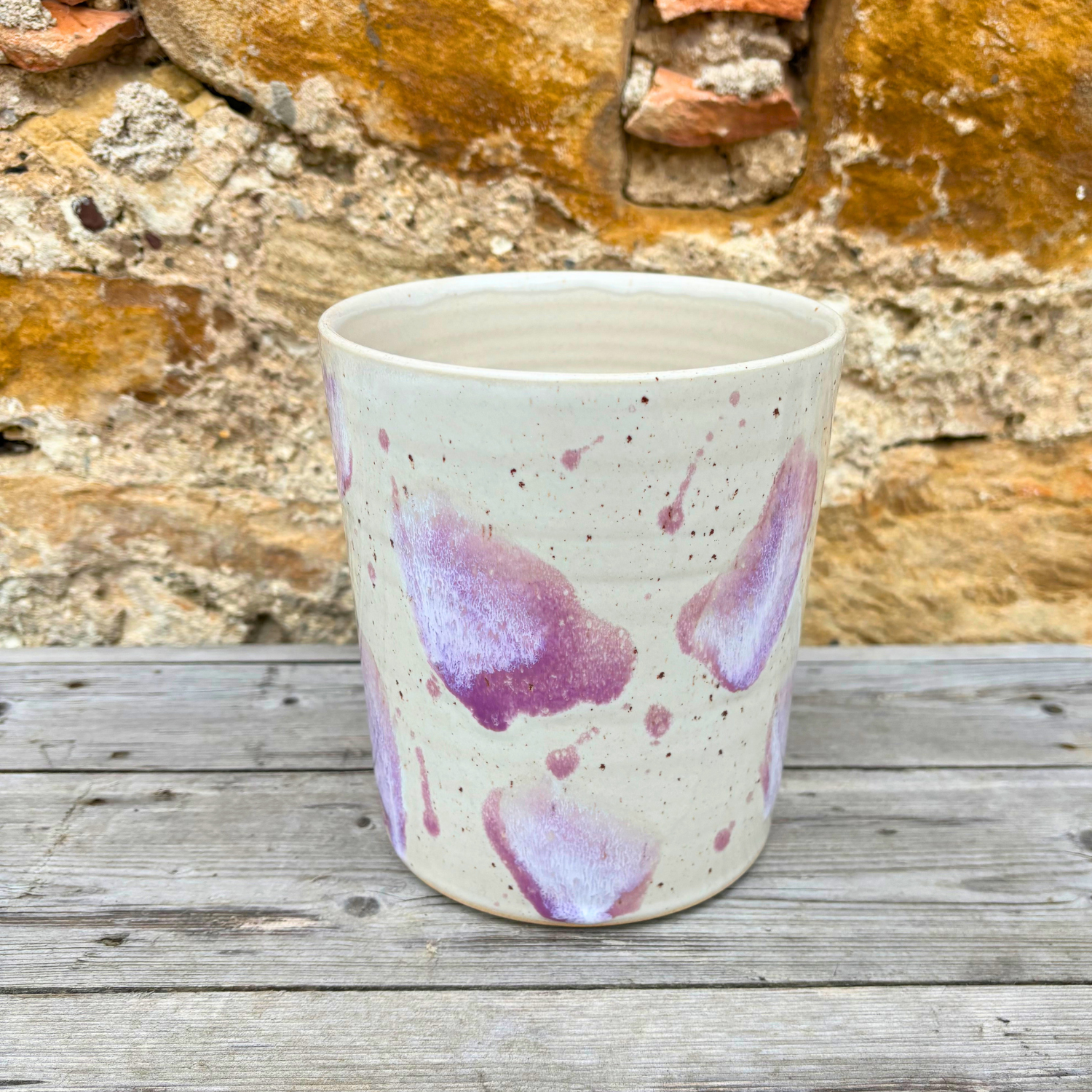 Ceramic utensil pot with purple speckles on a wooden surface with stone wall background