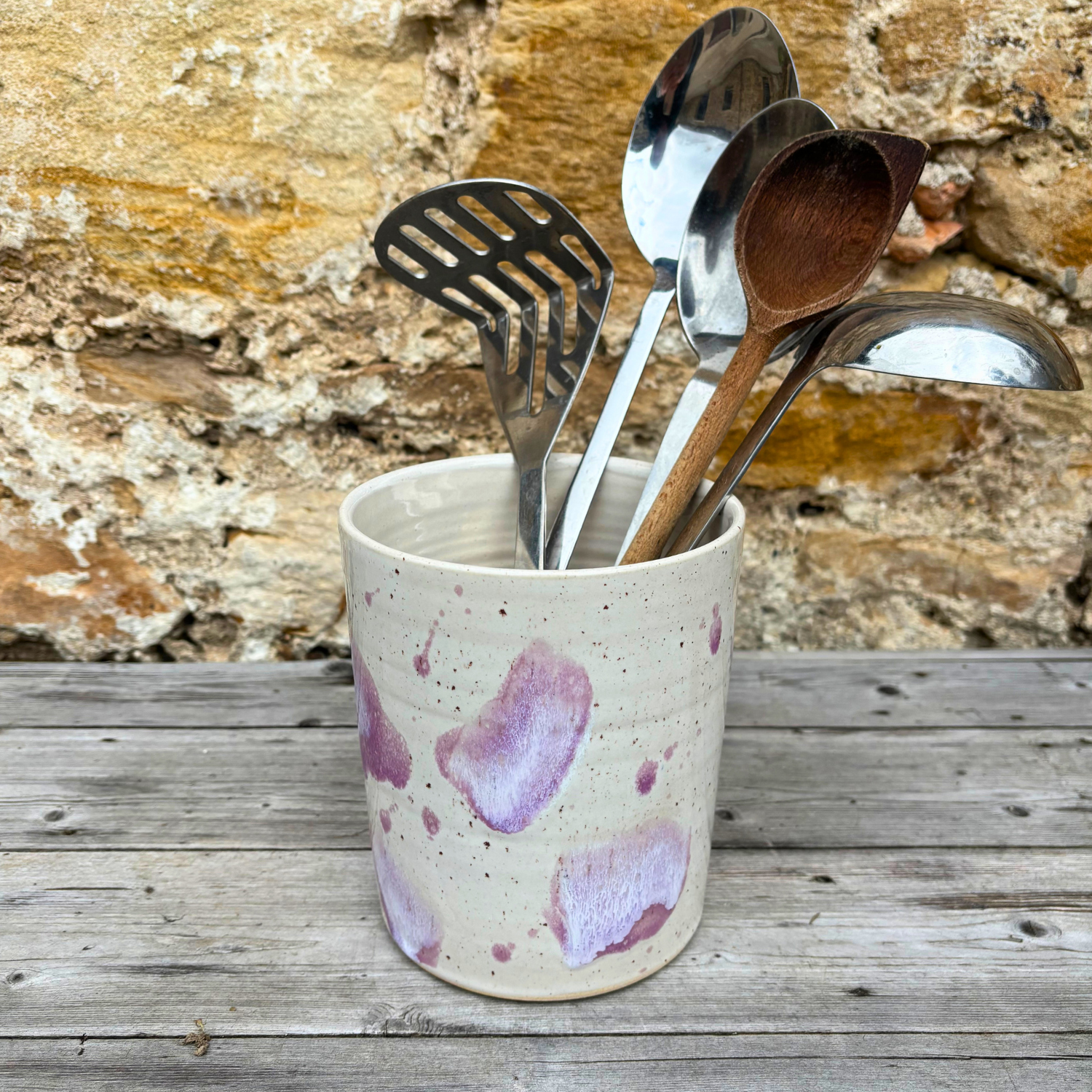 Ceramic container with pink speckles holding kitchen utensils against a stone wall background