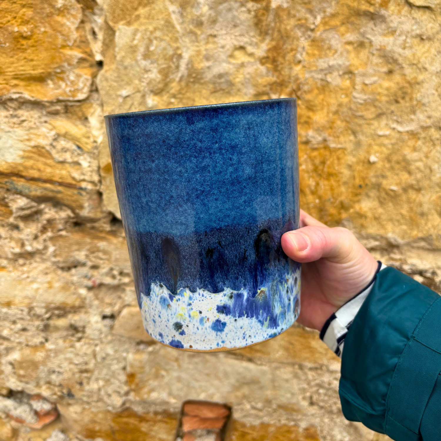 Hand holding a blue ceramic utensil pot against a stone wall background