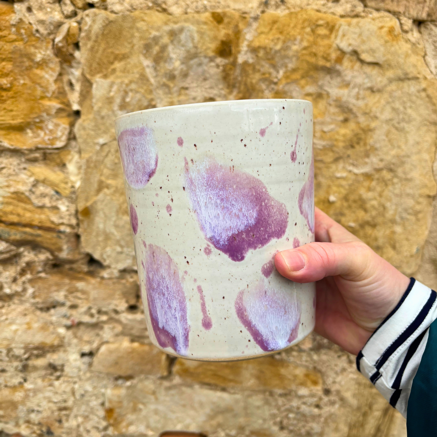 Hand holding a ceramic utensil pot with purple splashes against a stone wall background
