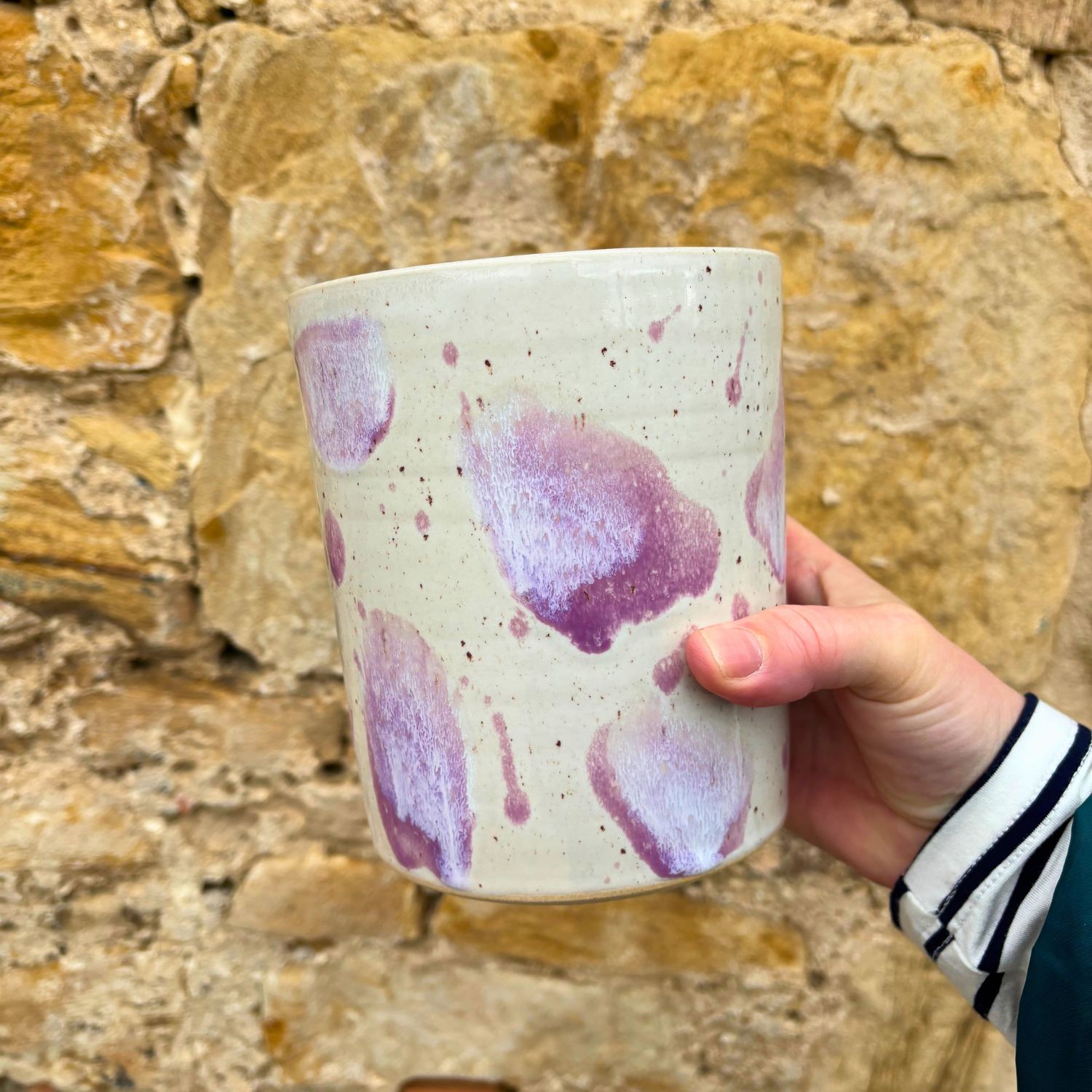 Hand holding a ceramic utensil pot with purple splashes against a stone wall background