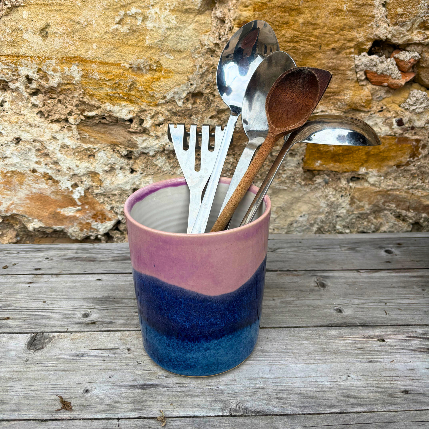 Pink and blue ceramic utensil holder with kitchen utensils against a stone wall background