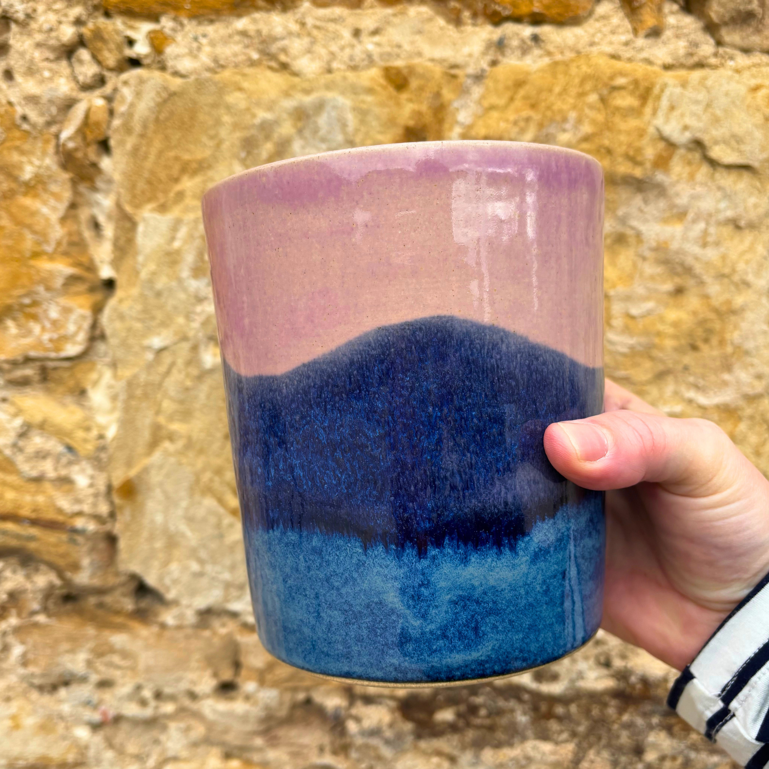 Hand holding a ceramic utensil pot with pink and blue gradient against a stone wall background