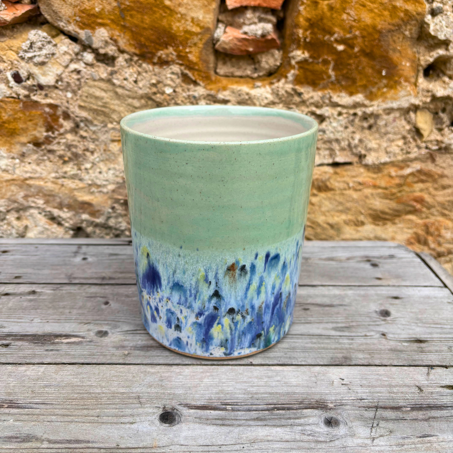Green ceramic pot with blue speckled design on a wooden surface with stone wall background