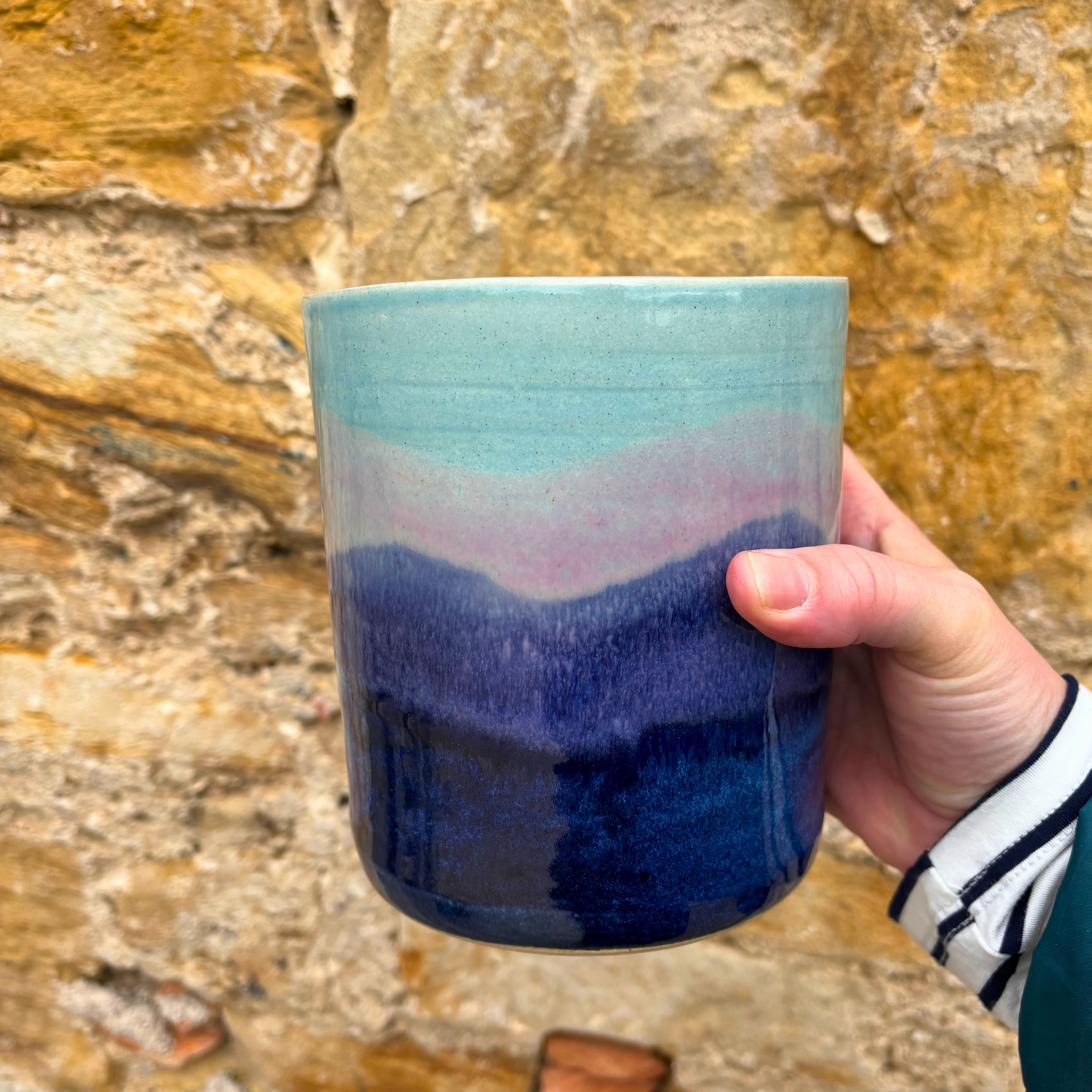 Hand holding a ceramic utensil pot with a gradient design against a stone wall background