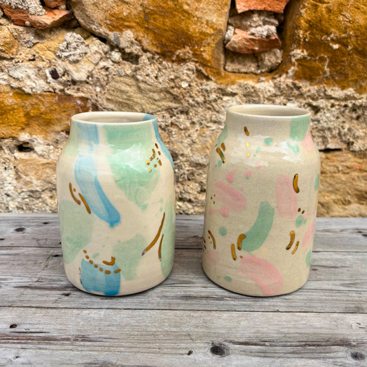 Two ceramic vases with abstract pastel designs on a wooden surface with a stone wall background.