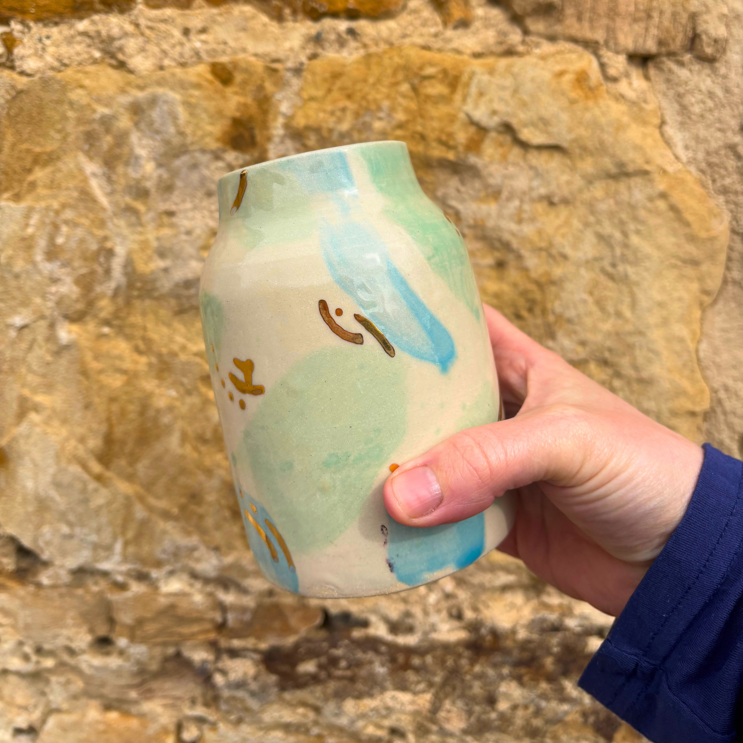 Hand holding a ceramic mug with artistic design against a stone wall background
