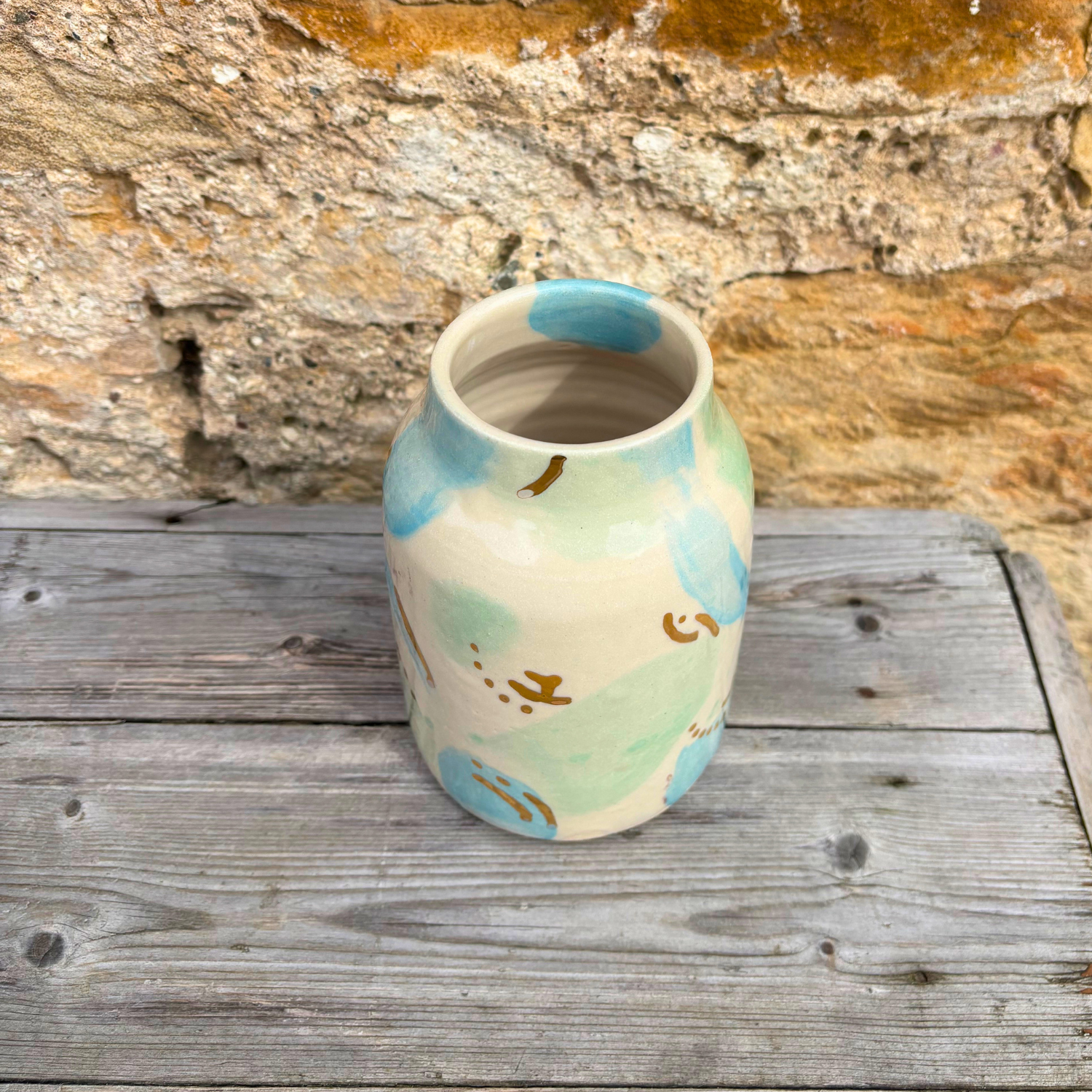 Ceramic vase with abstract design on a wooden surface against a stone wall.