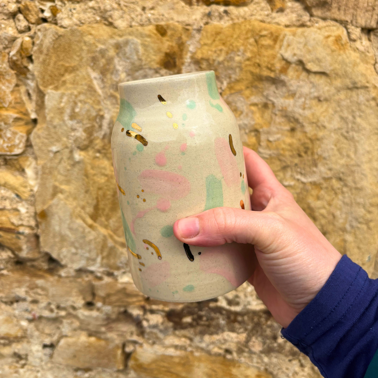 Hand holding a ceramic tumbler with colorful designs against a stone wall background