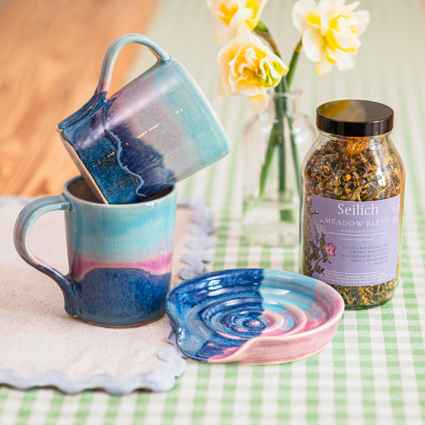 Two ceramic mugs and a spoon rest with a marbled design, alongside a jar of Seilich Meadow Blend tea on a checkered tablecloth.