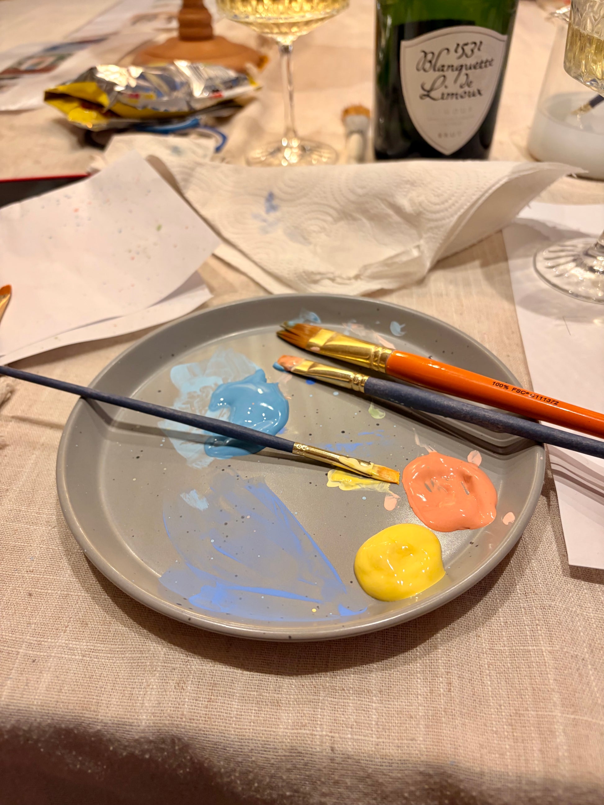 Paint palette with paintbrushes and color swatches on a table with a bottle of champagne in the background.