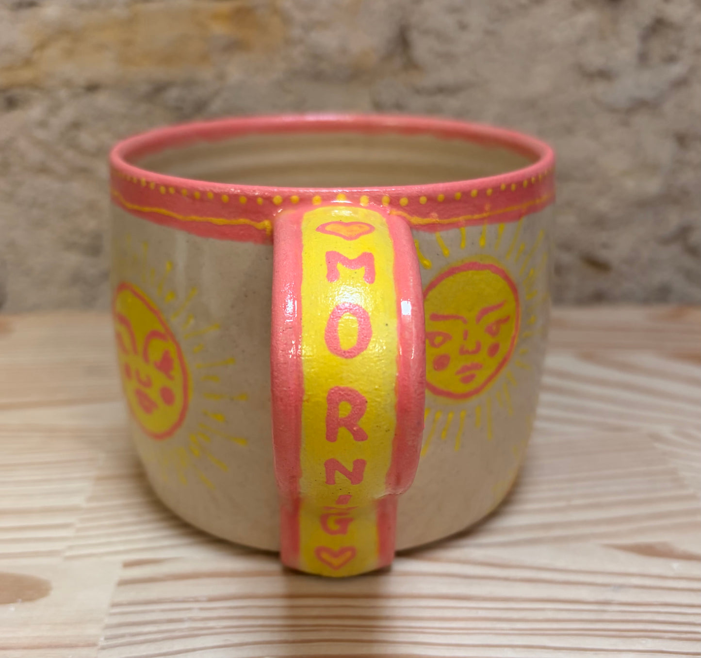 Ceramic mug with pink rim and yellow band reading 'MORNING' on a wooden surface.